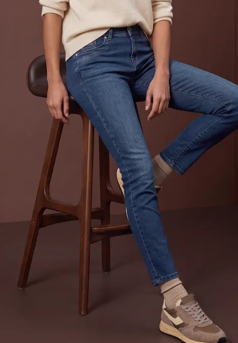 Mid Waist Straight Leg Jeans w kroju Slim Fit dark blue soft wash Osoba siedzi na drewnianym stołku, ma na sobie obcisłe niebieskie dżinsy i beżowe trampki.