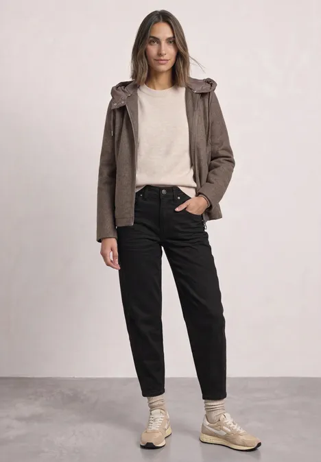 Frau in legerem Outfit mit brauner Jacke, hellem Pullover, schwarzer Hose und beigen Sneakern vor neutralem Hintergrund.