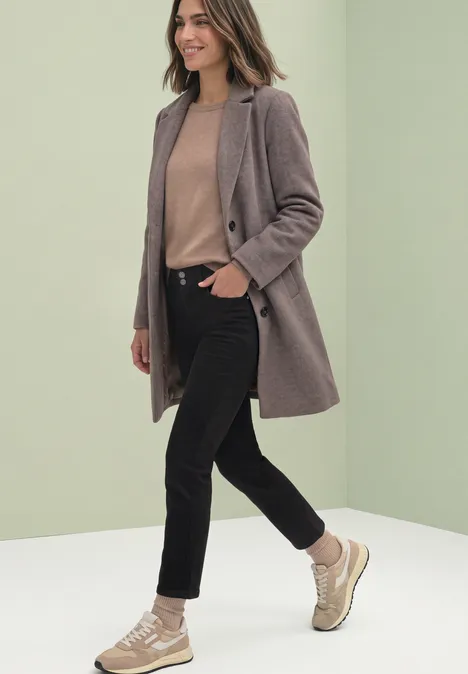 Mid Waist Straight Leg Jeans im Slim Fit soft black wash Frau in modernem Mantel, beigem Pullover, schwarzen Jeans und Sneakers vor hellgrünem Hintergrund.
