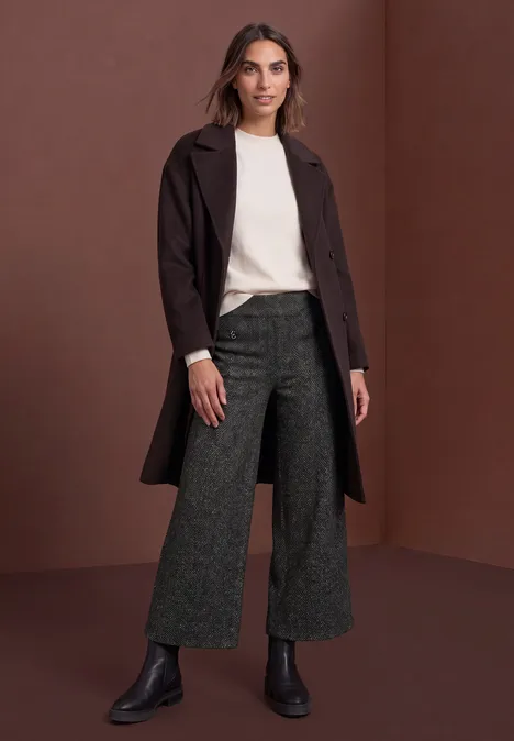 Frau in cremefarbenem Pullover, dunkelbraunem Mantel, weiten grauen Hosen und schwarzen Boots vor braunem Hintergrund.