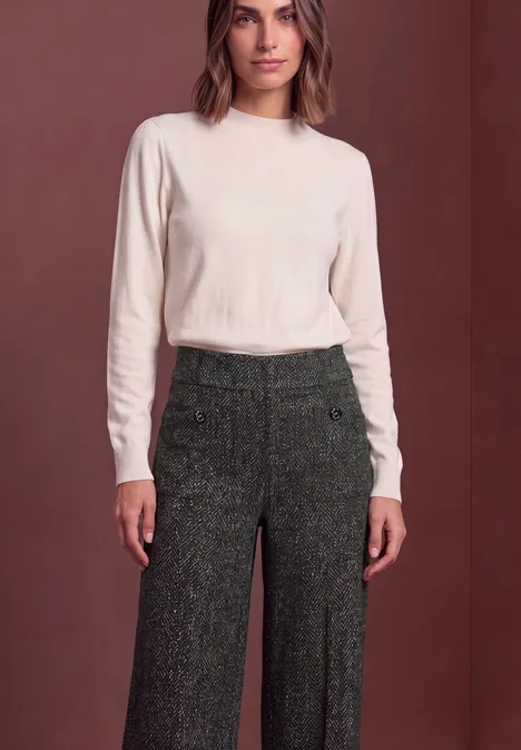 Frau in cremefarbenem Langarmshirt und eleganter, weiter, dunkelgrüner Tweed-Hose vor schlichtem Hintergrund.