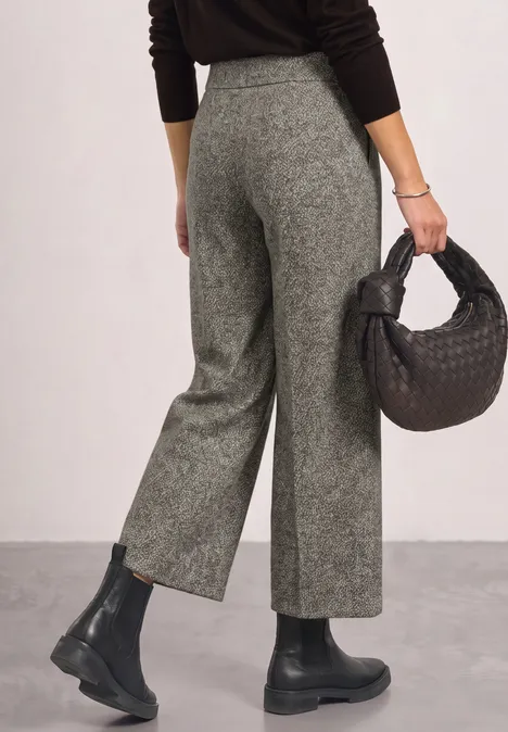 Frau trägt graue, hochgeschnittene Hose, schwarzen Pullover und Boots, hält eine schwarze geflochtene Tasche.