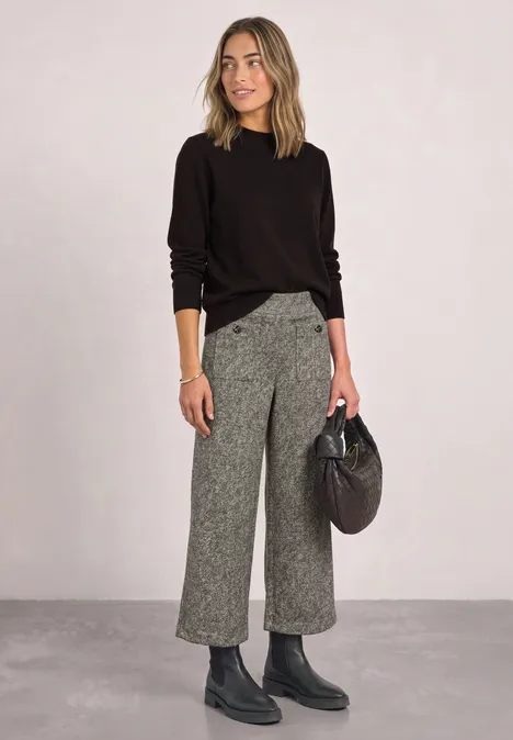 Frau in schwarzem Pullover, grauer Culotte, Boots und mit schwarzer Handtasche vor schlichtem Hintergrund.