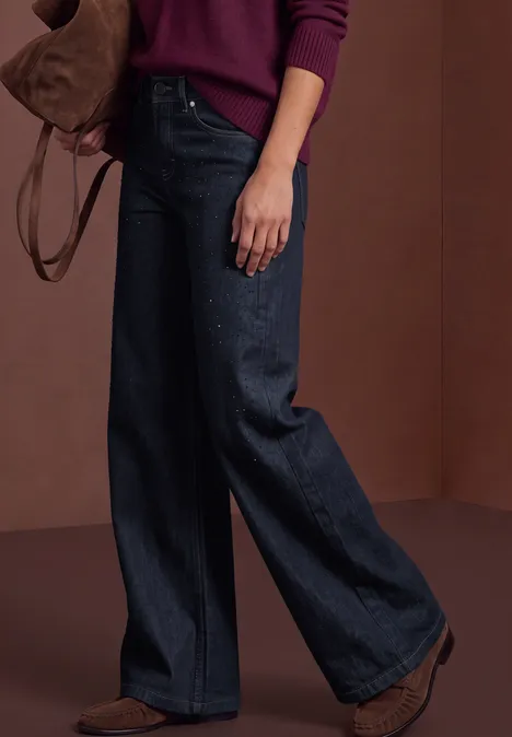 Frau trägt breite, dunkle Jeans mit braunen Stiefeln und einem Schulterbeutel, kombiniert mit einem kastanienroten Pullover.