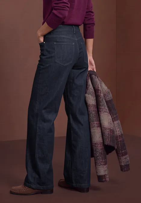Eine von hinten fotografierte Frau trägt dunkle, weite Jeans, braune Schuhe und hält eine karierte Jacke.