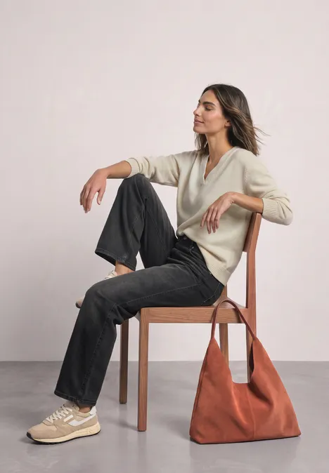 Een vrouw zit met gekruiste benen op een houten stoel, draagt een beige top, zwarte broek, sneakers en een rode tas.