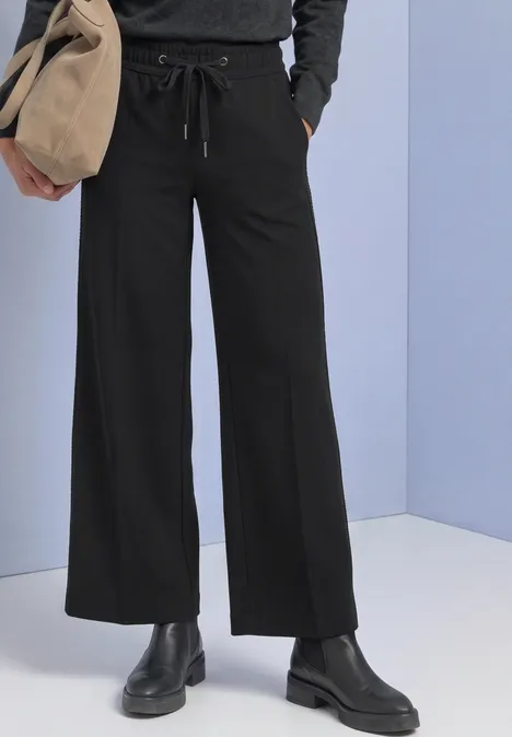 Zwarte, wijdvallende broek met aantrekkoord, zwarte laarzen en beige tas voor een lichtblauwe achtergrond.
