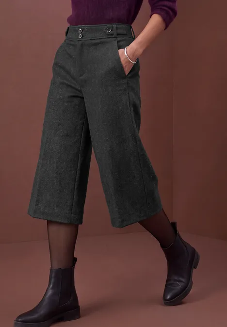 Pantalon culotte gris foncé, bottines noires et collants transparents sur fond rouge brique.