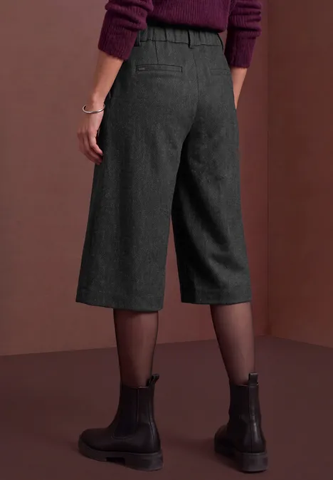 Vue arrière d’une personne portant un pantalon culotte noir, des collants transparents et des boots noires sur fond marron.