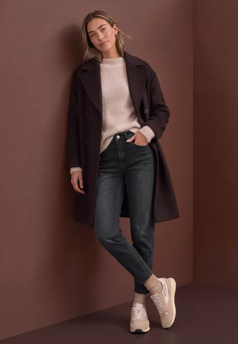 Jean taille haute à Barrel Leg, coupe ample grey washed Frau lehnt lässig an brauner Wand, trägt braunen Mantel, beigen Pullover, schwarze Jeans und helle Sneaker.