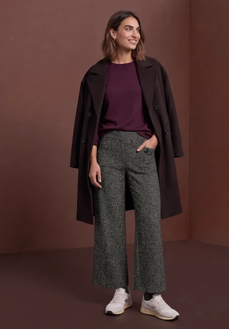 Frau trägt weinroten Pullover, dunklen Mantel, graue Culottes und weiße Sneaker vor schlichter brauner Wand.