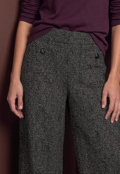 Ansicht von Taille bis Oberschenkeln in grauen, weiten Stoffhosen mit Knöpfen und lila Oberteil.