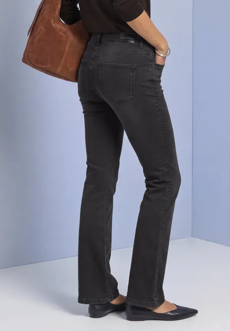 Frau trägt schwarze Jeans, dunkelblaue Ballerinas und eine braune Tasche vor hellblauem Hintergrund.