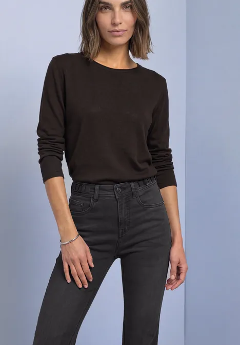 Frau in braunem Langarmshirt und dunkler Jeans steht vor neutralem Hintergrund.