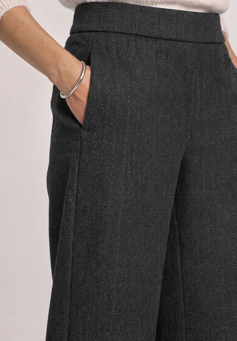 Pantalon en tissu gris foncé ample avec poches, main dans la poche, bracelet argenté visible.