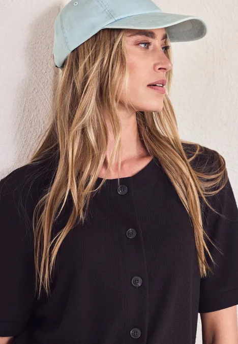 Personne portant une casquette, cheveux longs et haut noir à boutons, visage flou.