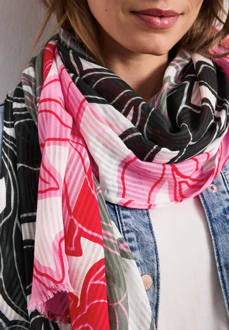 Frau mit pink-schwarzem, gemustertem Schal und Jeansjacke vor weißer Wand.