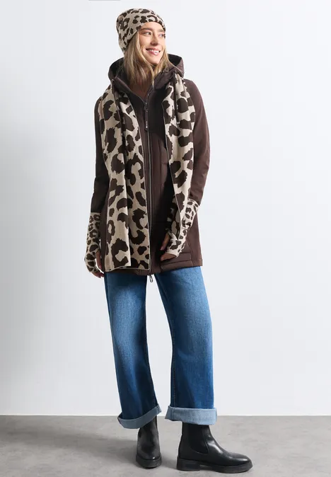 Eine Frau im Leoparden-Mützen-Set mit Schal und Handschuhen, braunem Mantel, weiter Jeans und schwarzen Boots.