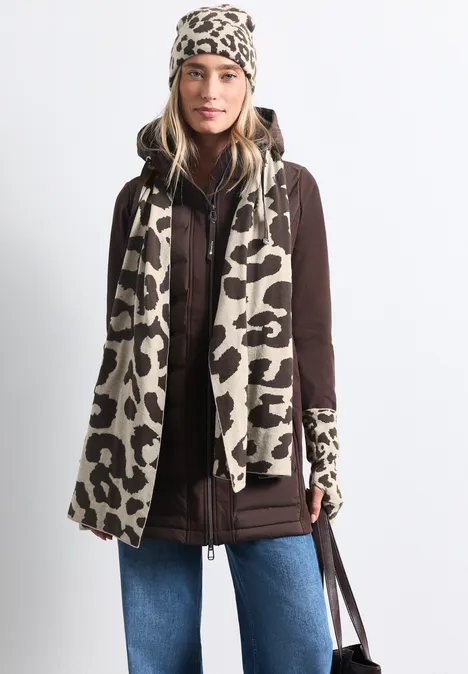 Frau trägt braune Jacke, blaue Jeans und beiges Leopardenmuster-Set mit Mütze, Schal und Handschuhen.