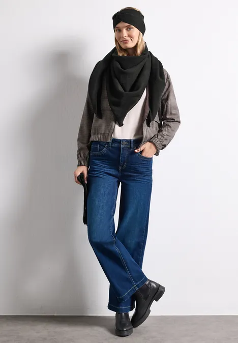 Vrouw met zwarte muts, driekhoekige sjaal, grijze jas, wit T-shirt, wijde jeans en zwarte laarzen.