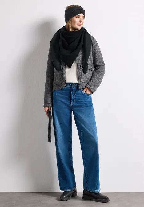 Frau in schwarzer Mütze und Schal, grauer Jacke, beigem Shirt, weiten Jeans und schwarzen Schuhen.