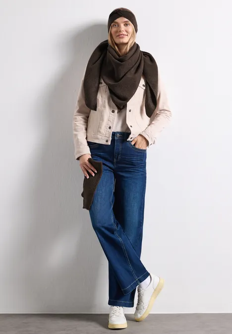 Glimlachend model in jeans, beige jasje, donkere sjaal en witte sneakers poseert voor een witte muur.