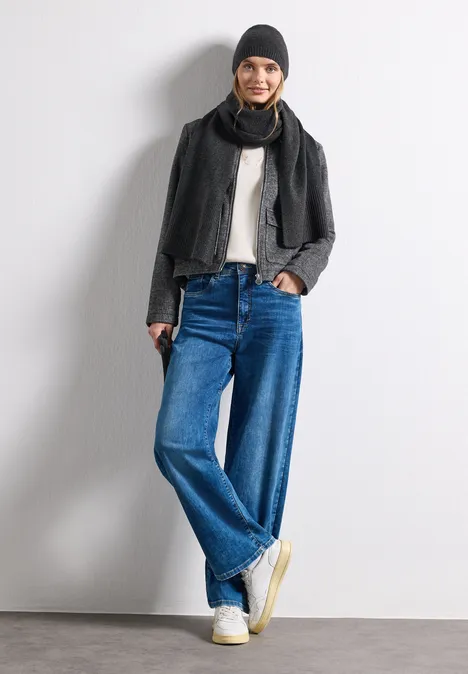 Vrouw in wijde jeans, wit shirt, zwarte jas, sjaal en muts, poseert ontspannen voor een witte achtergrond.