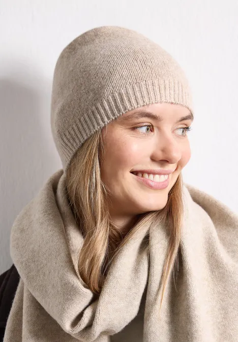 Feinstrick Beanie in Unifarbe Soft Sand Mel. Eine lächelnde Frau trägt eine beige Mütze und einen passenden Schal vor weißem Hintergrund.