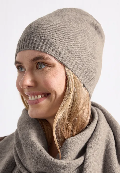 Feinstrick Beanie in Unifarbe Sandy Mocca Mel. Junge Frau lächelt seitlich, trägt graubeige Strickmütze und passenden Schal.