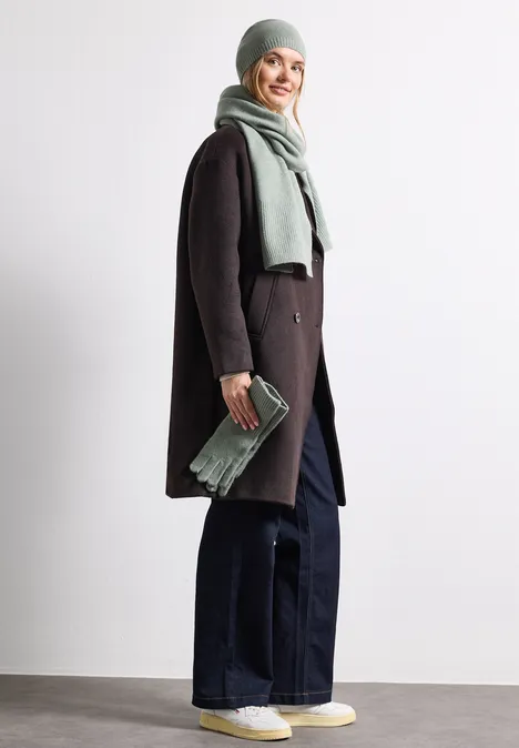 Feinstrick Beanie in Unifarbe eucalyptus green mel. Frau mit Wintermantel, Schal und Mütze, weiten Jeans und Sneakern, lächelnd und hält Clutch