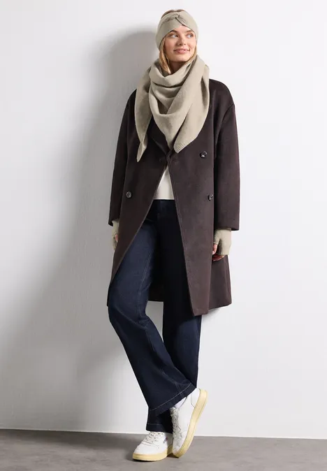 Fijn gebreide driehoekige sjaal in effen kleur Soft Sand Mel. Vrouw lacht in een donkere jas met beige sjaal, jeans en sneakers tegen een witte achtergrond.