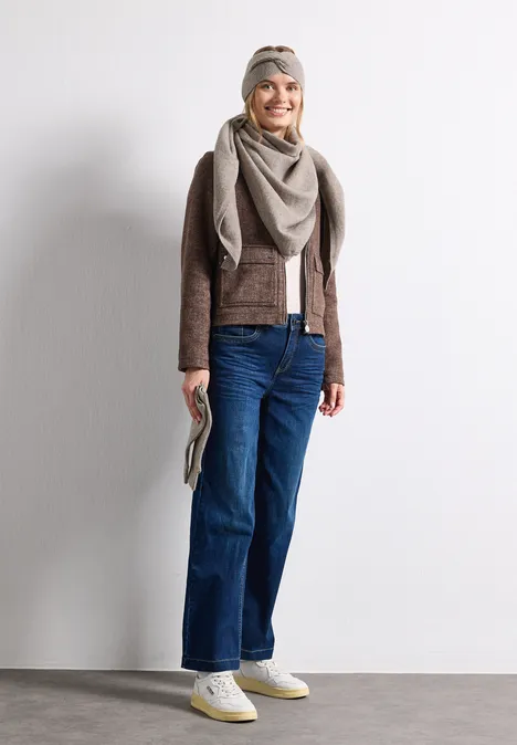 Fijn gebreide driehoekige sjaal in effen kleur Sandy Mocca Mel. Vrouw voor lichte muur, lacht, draagt jeans, bruine jas, sjaal, hoofddoek, witte sneakers en clutch.