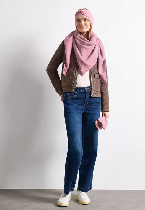Een vrouw draagt een bruine jas, blauwe spijkerbroek, roze sjaal en muts en houdt een roze handtas vast.