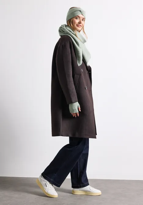 Fijn gebreide driehoekige sjaal in effen kleur eucalyptus green mel. Een glimlachende vrouw draagt een donkere jas, lichtgroene sjaal en muts, ruime broek en witte sneakers.