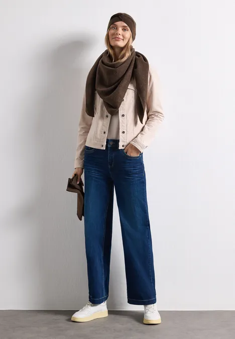Fijn gebreide driehoekige sjaal in effen kleur auburn brown mel. Jonge vrouw lacht, draagt muts, sjaal, lichte jas, wijde jeans en witte sneakers.