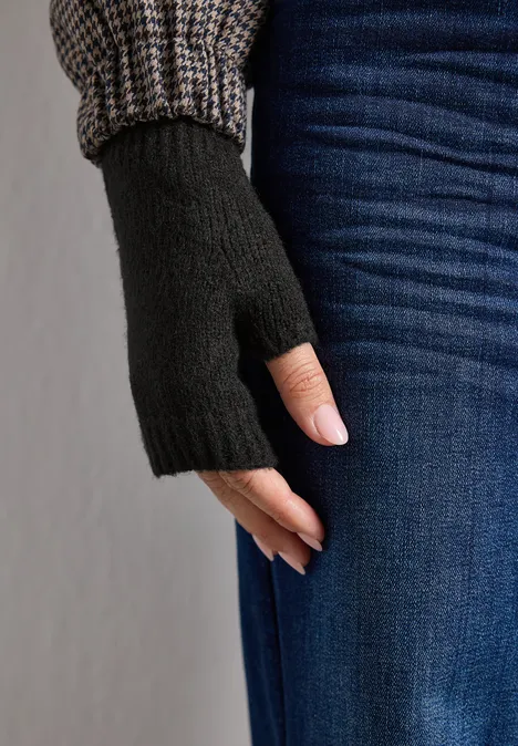 Schwarzer fingerloser Handschuh an einer Hand, kombiniert mit Jeans und Strickärmel im Hintergrund.