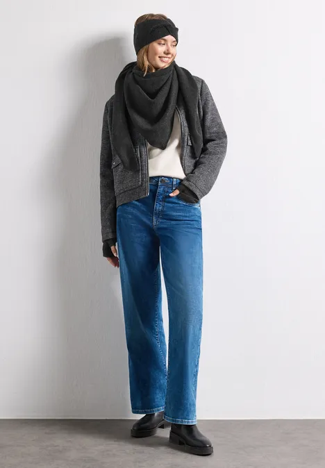 Frau lehnt an weißer Wand, trägt Beanie, Schal, schwarze Jacke, weiße Bluse und weite blaue Jeans mit schwarzen Schuhen.