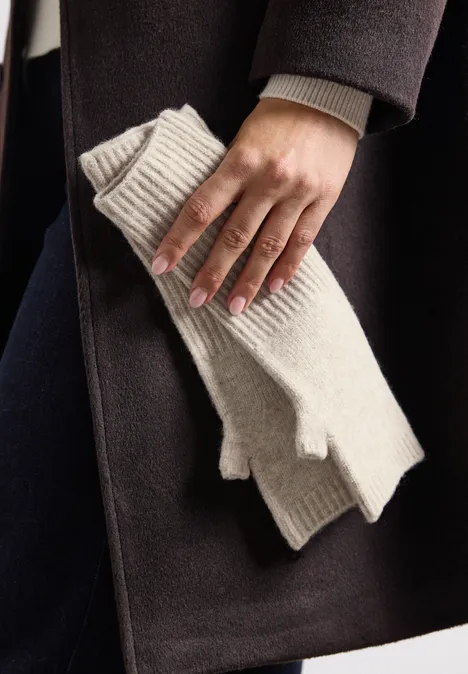 Een hand houdt crèmekleurige vingerloze gebreide wanten voor een donkere jas.