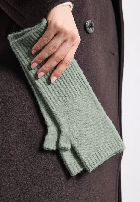 Manchettes en maille fine, couleur unie eucalyptus green mel. Eine Hand mit grünen, gerippten, fingerlosen Handschuhen vor einem dunklen Mantel