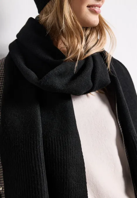 Une personne porte une écharpe noire en laine et un bonnet noir sur un pull beige clair.