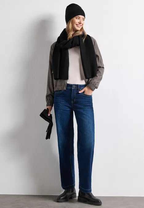 Femme en jean bleu, chemise claire, écharpe et bonnet noirs, mocassins, tenant un petit sac devant un mur gris