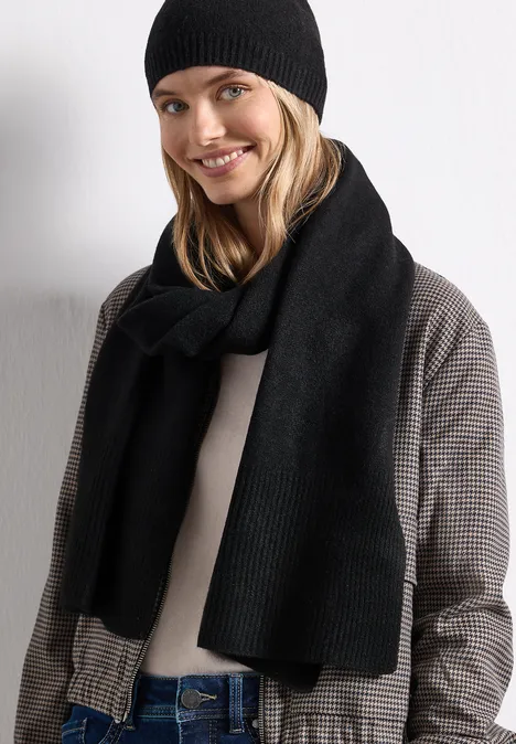 Jeune femme blonde souriant, porte un bonnet noir, une écharpe noire et une veste à carreaux devant un fond blanc.