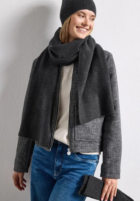 Frau mit dunkler Mütze, langer schwarzer Schal, grauer Jacke, beigem Pullover, Jeans und Clutch vor weißem Hintergrund.
