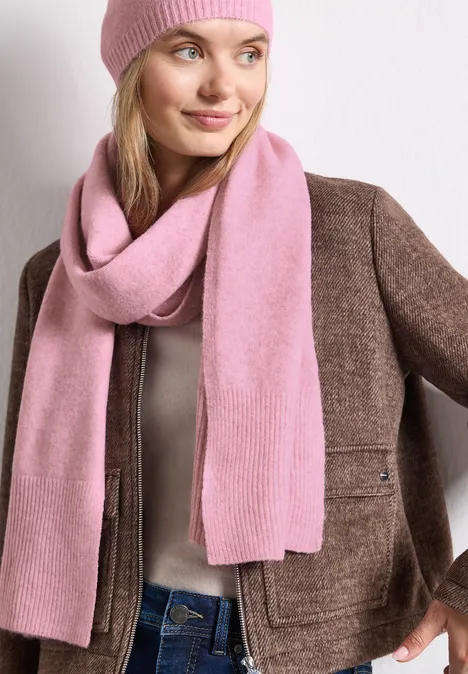 Langer Feinstrickschal in Unifarbe vintage blush mel. Frau mit rosa Mütze, rosa Schal und brauner Jacke vor hellem Hintergrund.