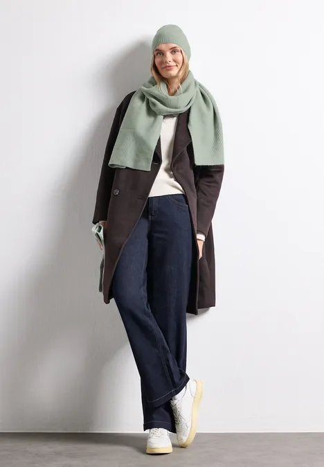 Femme avec bonnet et écharpe vert clair, manteau sombre, jean large et baskets blanches sur fond blanc.