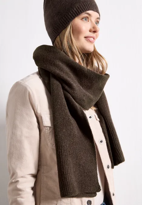 Langer Feinstrickschal in Unifarbe auburn brown mel. Frau mit brauner Strickmütze und Schal, helle Jacke, lächelnd seitlich blickend.