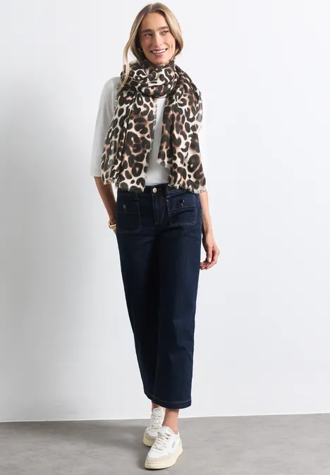 Frau in weißem Langarmshirt, Leopardenmuster-Schal, weiten Jeans und weißen Sneakers vor hellem Hintergrund.