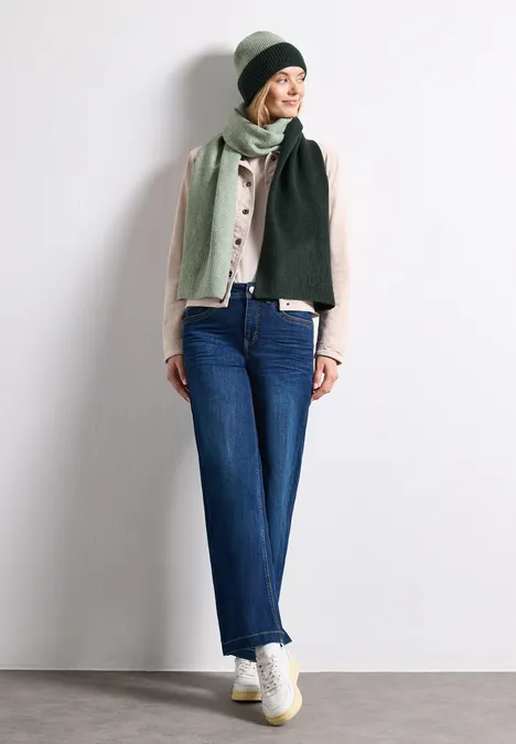 Écharpe en maille fine au look « color block » mossbound green Frau mit beigem Hut, zweifarbigem grünem Schal, Bluse, blauen Jeans und weißen Sneakern vor neutralem Hintergrund.