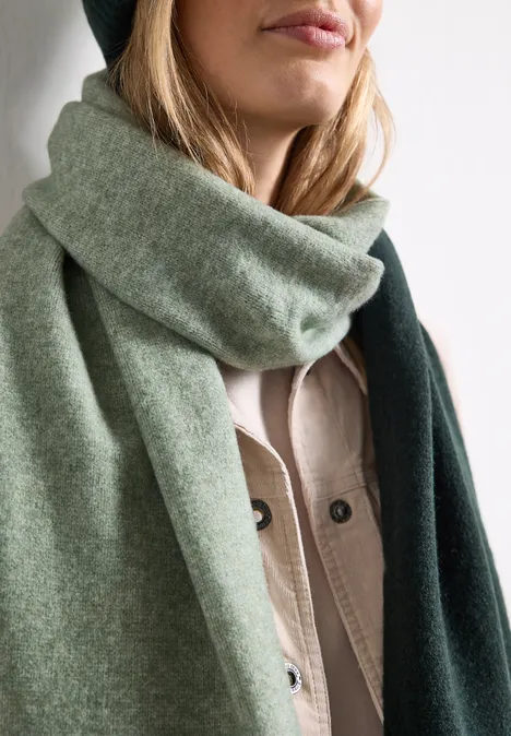 Écharpe en maille fine au look « color block » mossbound green Frau mit hellgrün-dunklem Schal um den Hals, trägt beigefarbene Jacke, vor weißem Hintergrund.