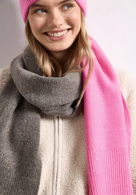 Junge Frau lächelt, trägt pinke Mütze und zweifarbigen Wollschal (pink und grau) sowie helle Jacke.